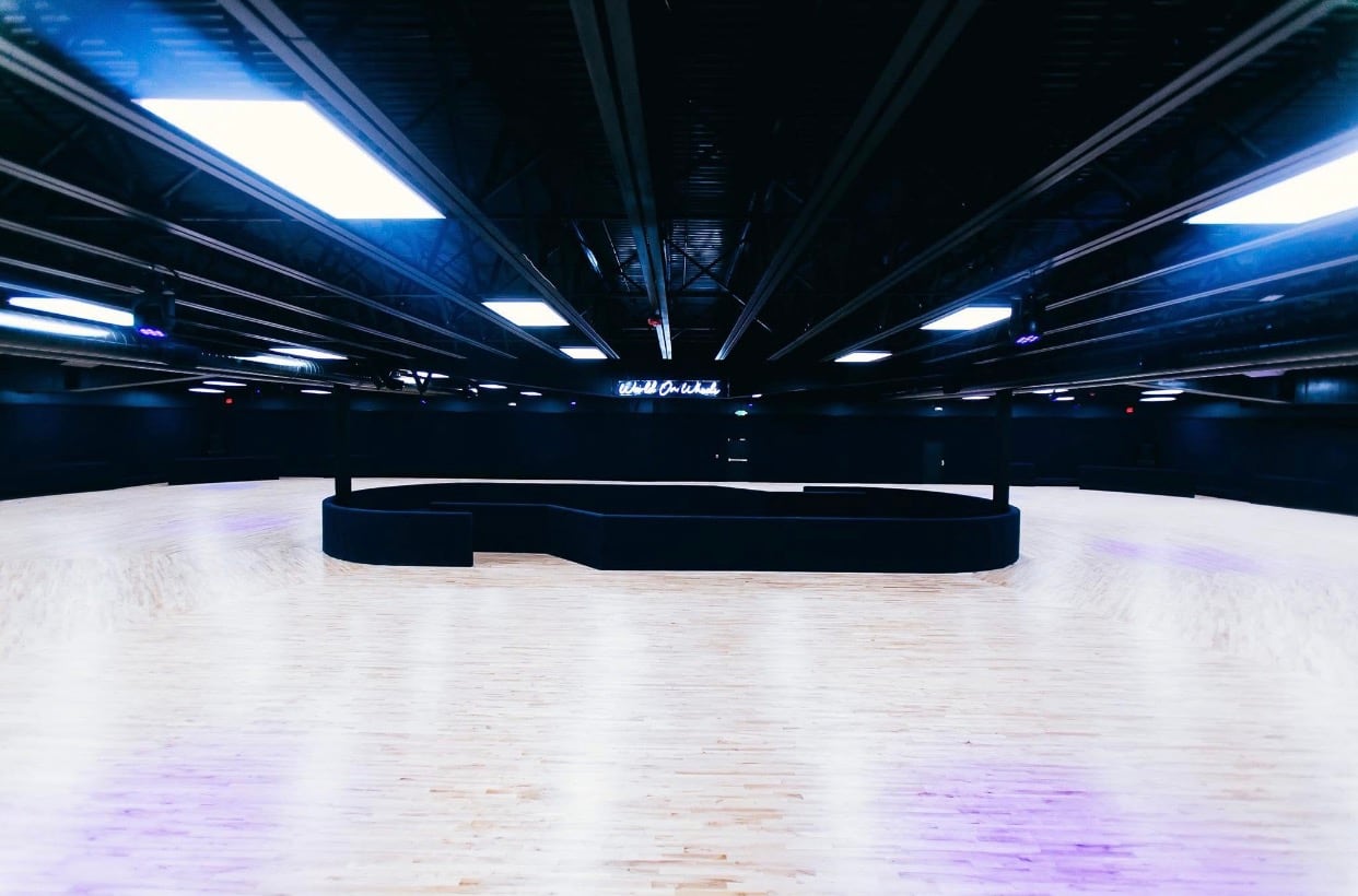 The empty maple skating floor at World on Wheels under dark trusses and bright panel lights, with the neon World On Wheels sign glowing on the far wall