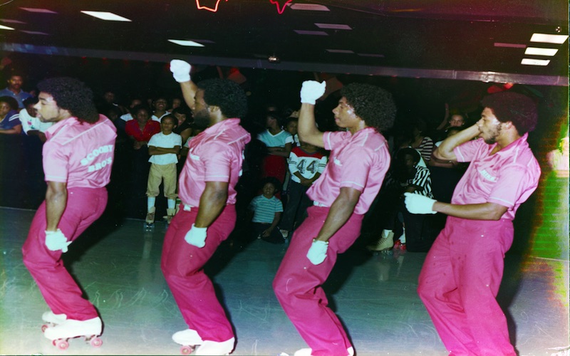Four men in matching pink outfits and white gloves skate in step on a rink floor while a crowd watches from the edge